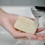 Unrecognizable person standing near sink with soap in hand under water pouring from tap during daily hygienic procedure in bathroom